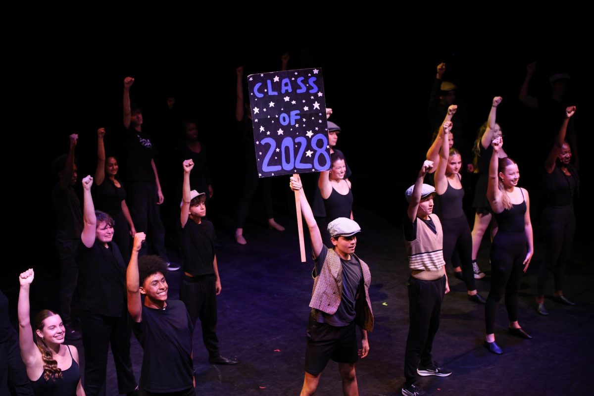 Raising a sign in the air reading “Class of 2028,” theatre freshman Tristan Dominguez performs in a group piece titled, “Seize the Day” from “Newsies” by Alan Menken.